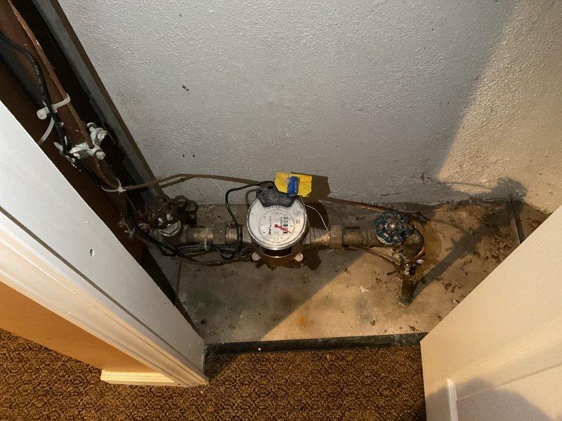 Responded to severe leak at exterior faucet mounted on back wall beneath kitchen window where water infiltration had begun entering the home and causing dirt accumulation on interior windowsill. Inspected outdoor spigot installation on 60-year-old brick structure near generator and assessed water meter assembly in basement access area showing significant staining on concrete base, indicating chronic moisture issues. Documented exterior wall conditions including utility connections, PVC drainage, glass block basement window, and foundation plantings, as well as interior meter installation with copper supply lines and shut-off valves to determine source and extent of leak for repair recommendations.
