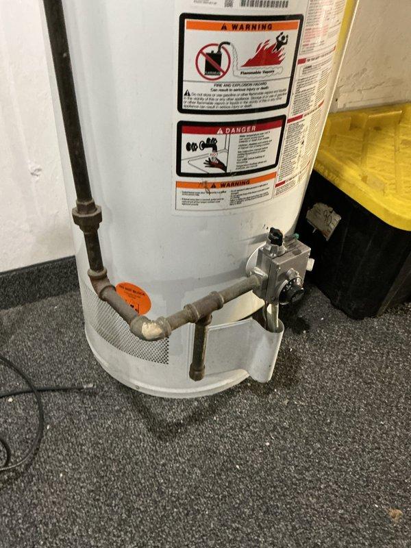 Completed stack repair and water heater installation with associated plumbing connections. Installed new water heater unit on carpeted floor with proper drain pan for leak protection, connected copper supply lines and gas control valve assembly at base of unit. Verified all safety labels and warning placards properly affixed to exterior of tank. Configured underside mechanical connections including copper piping with threaded fittings, flexible braided lines, shutoff valve with red handle lever, and black canister component. All plumbing connections secured and tested for proper operation.