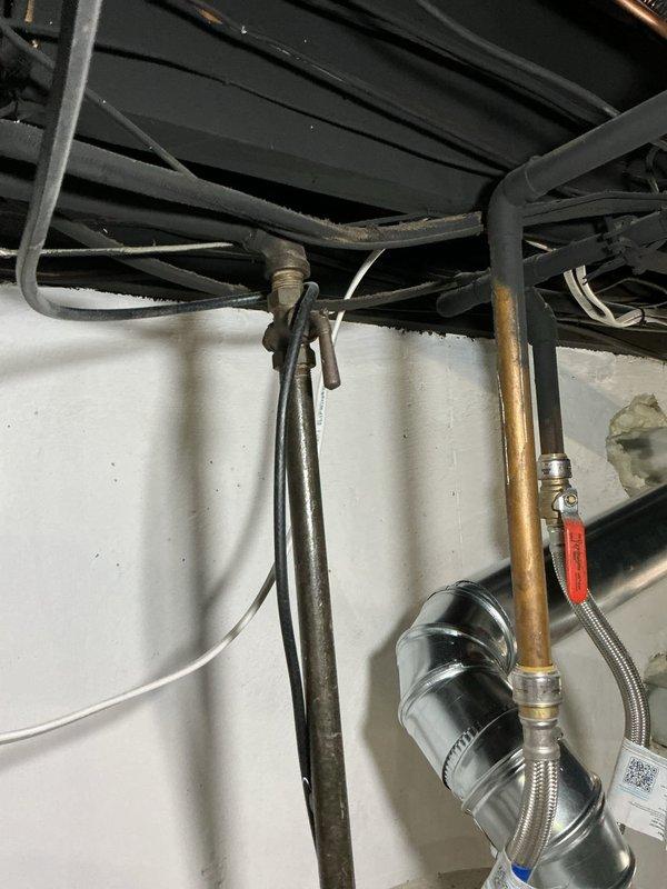Completed stack repair and water heater installation with associated plumbing connections. Installed new water heater unit on carpeted floor with proper drain pan for leak protection, connected copper supply lines and gas control valve assembly at base of unit. Verified all safety labels and warning placards properly affixed to exterior of tank. Configured underside mechanical connections including copper piping with threaded fittings, flexible braided lines, shutoff valve with red handle lever, and black canister component. All plumbing connections secured and tested for proper operation.