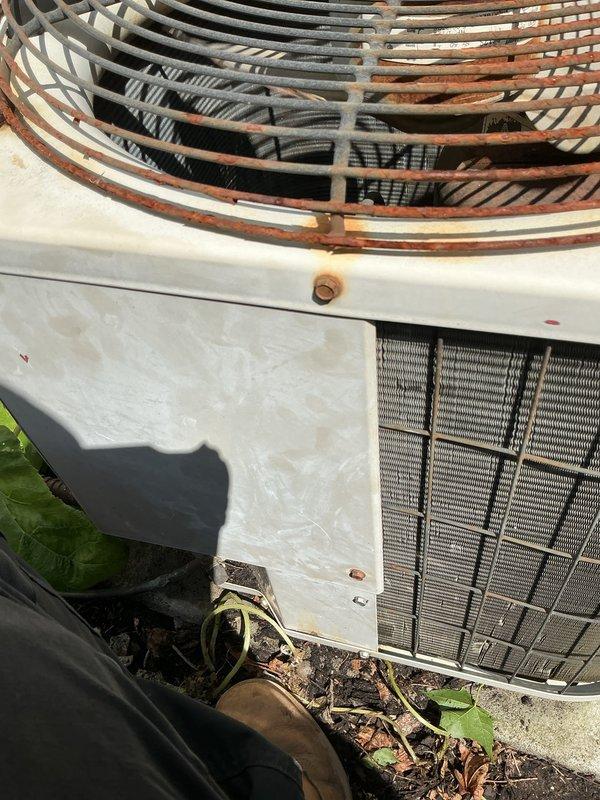 Completed comprehensive Gold AC Inspection and tune-up on residential split system with 5-year-old outdoor condensing unit featuring a 34-year-old compressor. Performed visual inspection of condenser unit positioned on concrete pad at rear of property, noting significant rust accumulation on fan guard and general weathering consistent with age. Cleared debris and leaves from base of unit and surrounding area. Inspected condenser coils through protective grille, verified electrical disconnect box and conduit connections at wall mount. Checked fan guard integrity and internal components visibility. Unit operational at time of inspection with no reported symptoms or performance issues prior to service call. Documented equipment condition through photographic records showing exterior cabinet, top-mounted fan assembly with radial guard pattern, and positioning relative to structure with vinyl siding. System demonstrated typical wear patterns expected for equipment age, particularly on expos