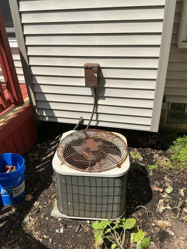 Completed comprehensive Gold AC Inspection and tune-up on residential split system with 5-year-old outdoor condensing unit featuring a 34-year-old compressor. Performed visual inspection of condenser unit positioned on concrete pad at rear of property, noting significant rust accumulation on fan guard and general weathering consistent with age. Cleared debris and leaves from base of unit and surrounding area. Inspected condenser coils through protective grille, verified electrical disconnect box and conduit connections at wall mount. Checked fan guard integrity and internal components visibility. Unit operational at time of inspection with no reported symptoms or performance issues prior to service call. Documented equipment condition through photographic records showing exterior cabinet, top-mounted fan assembly with radial guard pattern, and positioning relative to structure with vinyl siding. System demonstrated typical wear patterns expected for equipment age, particularly on expos