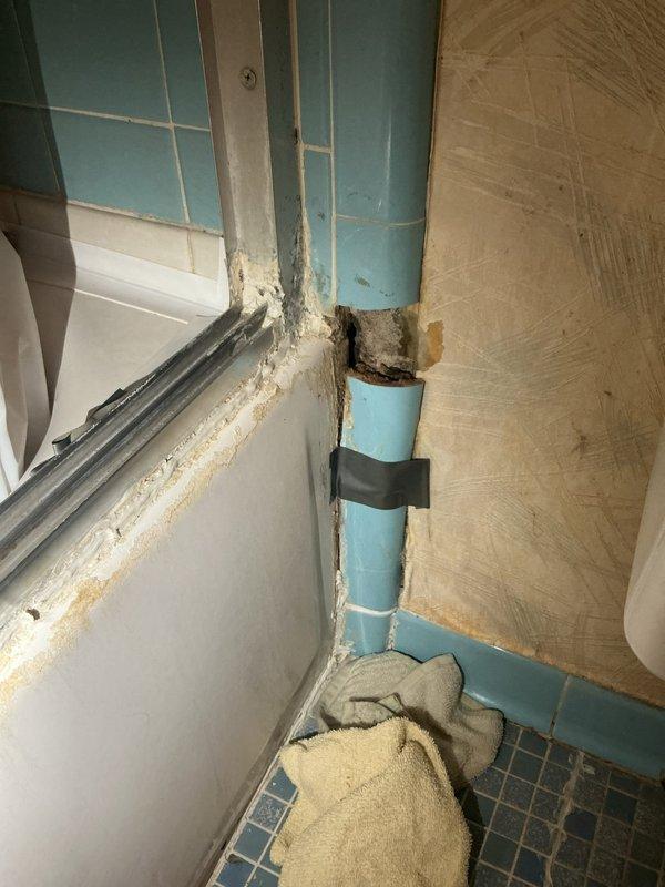Responded to callback for water leak originating from bathtub area affecting kitchen ceiling below. Upon inspection, discovered severely deteriorated grout and water damage around vertical PVC drain pipe in bathroom corner, with extensive mold growth and material degradation at wall and floor junction indicating chronic moisture intrusion. Investigation revealed active leak path from compromised bathroom plumbing penetrating through floor assembly to kitchen ceiling, evidenced by visible ceiling damage and access holes created for leak tracing. Documented conditions showing failed waterproofing at tile-to-pipe interface and degraded grout lines allowing water migration between floors.