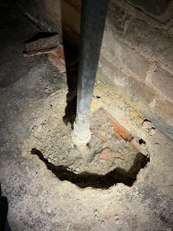 Responded to a cracked main water line located at the basement entry point on the main floor of a 40-year-old residence. Upon arrival, accessed the affected area where flooring had been opened to expose the damaged pipe, which had been leaking for approximately seven days. Conducted thorough inspection of the vertical galvanized steel water supply line and surrounding concrete foundation area, documenting significant deterioration around the pipe penetration point with visible concrete crumbling, exposed aggregate, and rust staining. Assessed the blue-handled shutoff valve and traced the water line along the basement wall, noting extensive water staining and moisture damage patterns on concrete surfaces indicating active leak history. Photographed the compromised pipe section, deteriorated foundation penetration, and water damage evidence for repair documentation. Confirmed damage was localized to the main floor entry area with no extension to other zones of the residence. Recommended 