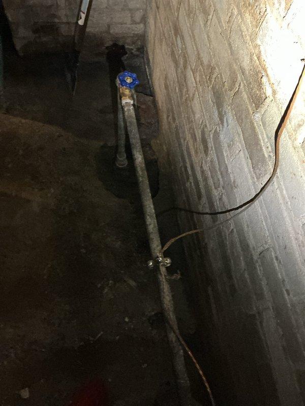 Responded to a cracked main water line located at the basement entry point on the main floor of a 40-year-old residence. Upon arrival, accessed the affected area where flooring had been opened to expose the damaged pipe, which had been leaking for approximately seven days. Conducted thorough inspection of the vertical galvanized steel water supply line and surrounding concrete foundation area, documenting significant deterioration around the pipe penetration point with visible concrete crumbling, exposed aggregate, and rust staining. Assessed the blue-handled shutoff valve and traced the water line along the basement wall, noting extensive water staining and moisture damage patterns on concrete surfaces indicating active leak history. Photographed the compromised pipe section, deteriorated foundation penetration, and water damage evidence for repair documentation. Confirmed damage was localized to the main floor entry area with no extension to other zones of the residence. Recommended 
