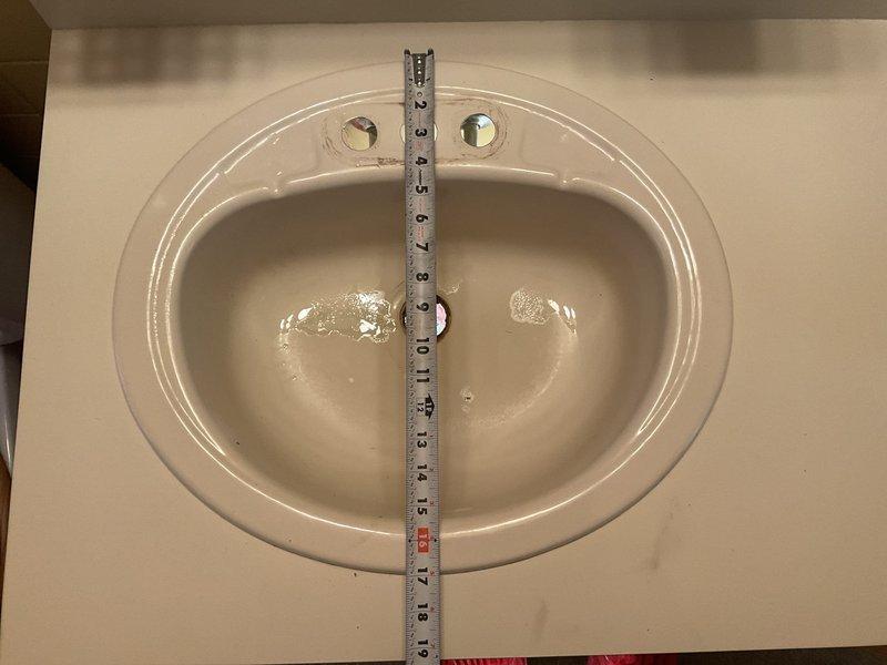 Performed bathroom sink measurement and assessment. Documented existing oval porcelain sink basin dimensions and faucet mounting configuration with three-hole setup for standard widespread or centerset installation. Captured detailed measurements of interior basin width and depth using precision measuring tools to ensure accurate specifications for potential replacement or repair work. Noted current condition of fixture including water staining and verified drain opening accessibility for future service needs.
