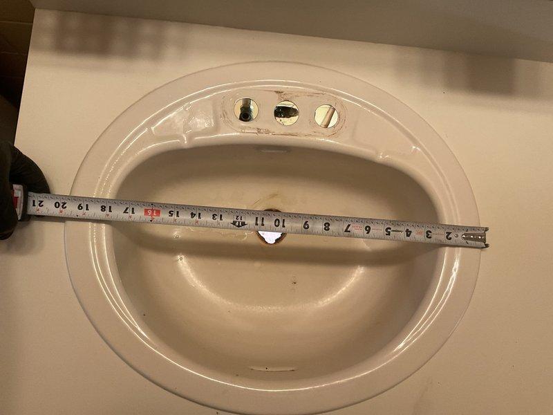 Performed bathroom sink measurement and assessment. Documented existing oval porcelain sink basin dimensions and faucet mounting configuration with three-hole setup for standard widespread or centerset installation. Captured detailed measurements of interior basin width and depth using precision measuring tools to ensure accurate specifications for potential replacement or repair work. Noted current condition of fixture including water staining and verified drain opening accessibility for future service needs.