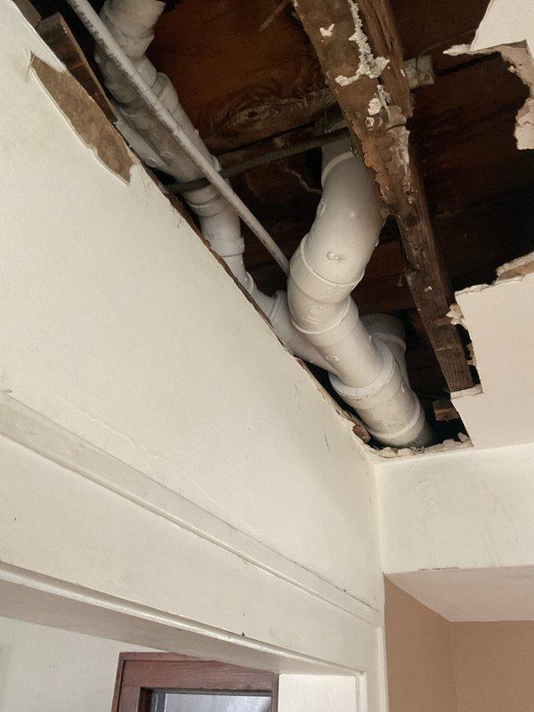 Completed toilet replacement and associated plumbing repair in residential bathroom. Removed existing toilet fixture and addressed drainage system issues requiring access through ceiling from floor above. Installed new white porcelain two-piece toilet with chrome flush handle in narrow bathroom alcove with tile flooring and wood trim. Repaired PVC drainage piping running through ceiling joists, including angled pipe runs and curved joints to ensure proper drainage flow. Patched and prepared ceiling opening for drywall restoration following plumbing repairs. All fixtures tested and verified operational upon completion.