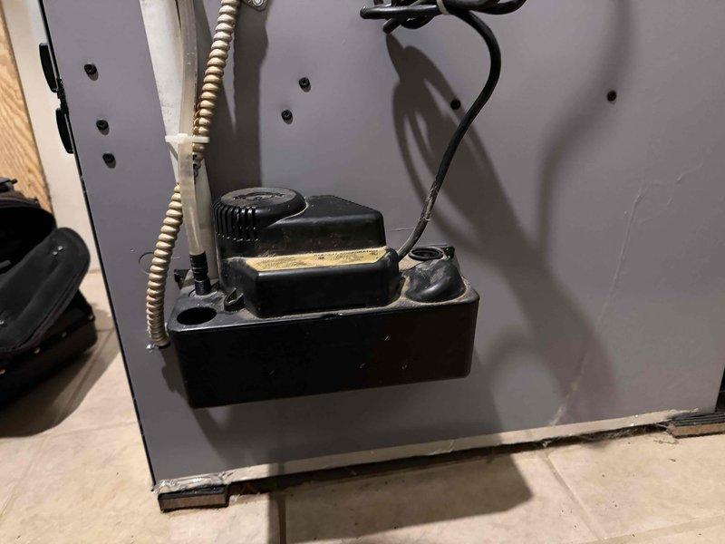 Responded to service call for condensate pump running dry and humming on 6-year-old Thermal Zone unit located in basement. Upon inspection, found condensate pump assembly mounted at base of equipment with flexible conduit and drainage lines routing upward from unit. Assessed pump operation, electrical connections, and drainage system configuration. Documented equipment condition and pump installation with photos showing black rectangular pump assembly with cylindrical motor housing, flexible metal conduit, and associated wiring connections mounted to gray HVAC panel.