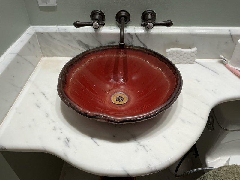 Completed installation of decorative vessel sink basin in bathroom with coordinating wall-mounted faucet assembly. Installed round bowl-style vessel sink with scalloped rim design onto existing white marble countertop with gray veining. Mounted three-piece wall-mounted faucet system in oil-rubbed bronze finish, consisting of two handle controls and center spout positioned above vessel basin. Connected water supply lines to wall-mounted valves and tested for proper flow and temperature control. Installed matching pop-up drain assembly with bronze finish drain cover in sink basin and verified proper drainage function. Secured all mounting hardware and sealed connections to prevent leaks. Performed final inspection to ensure all fixtures were properly aligned, fully operational, and aesthetically coordinated with existing bathroom finishes.
