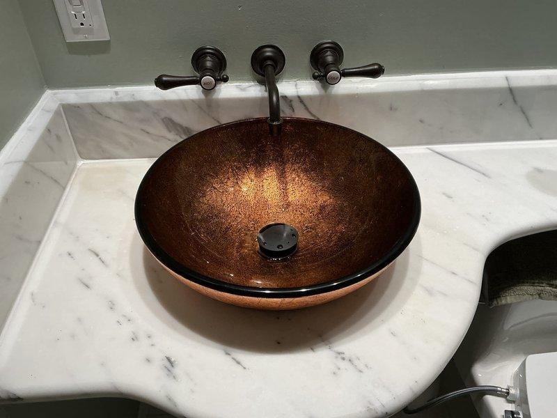 Completed installation of decorative vessel sink basin in bathroom with coordinating wall-mounted faucet assembly. Installed round bowl-style vessel sink with scalloped rim design onto existing white marble countertop with gray veining. Mounted three-piece wall-mounted faucet system in oil-rubbed bronze finish, consisting of two handle controls and center spout positioned above vessel basin. Connected water supply lines to wall-mounted valves and tested for proper flow and temperature control. Installed matching pop-up drain assembly with bronze finish drain cover in sink basin and verified proper drainage function. Secured all mounting hardware and sealed connections to prevent leaks. Performed final inspection to ensure all fixtures were properly aligned, fully operational, and aesthetically coordinated with existing bathroom finishes.