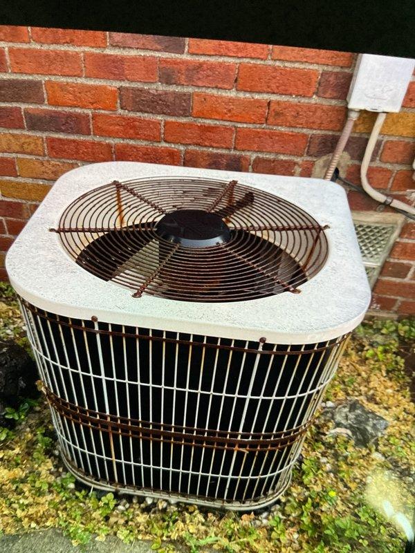 Completed comprehensive Gold AC inspection on 12-year-old single-unit residential air conditioning system with outdoor condenser unit located adjacent to building foundation and air handler positioned in basement. Performed thorough visual assessment of condenser unit exterior components, including top-mounted fan assembly with protective guard, vertical louvered side panels, and housing integrity. Inspected unit for signs of age-related wear, noting weathering and surface oxidation consistent with outdoor exposure on metal components and fan guard. Evaluated ground-level installation conditions, including gravel pad placement, drainage patterns, and clearance from brick and siding exterior walls. Verified adequate airflow access through intake grilles and assessed surrounding area for debris accumulation. Documented equipment condition through photographic records showing condenser positioning, component configuration, and overall operational setup. System confirmed operational at tim