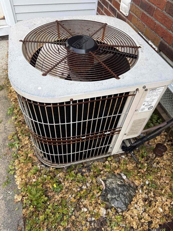 Completed comprehensive Gold AC inspection on 12-year-old single-unit residential air conditioning system with outdoor condenser unit located adjacent to building foundation and air handler positioned in basement. Performed thorough visual assessment of condenser unit exterior components, including top-mounted fan assembly with protective guard, vertical louvered side panels, and housing integrity. Inspected unit for signs of age-related wear, noting weathering and surface oxidation consistent with outdoor exposure on metal components and fan guard. Evaluated ground-level installation conditions, including gravel pad placement, drainage patterns, and clearance from brick and siding exterior walls. Verified adequate airflow access through intake grilles and assessed surrounding area for debris accumulation. Documented equipment condition through photographic records showing condenser positioning, component configuration, and overall operational setup. System confirmed operational at tim