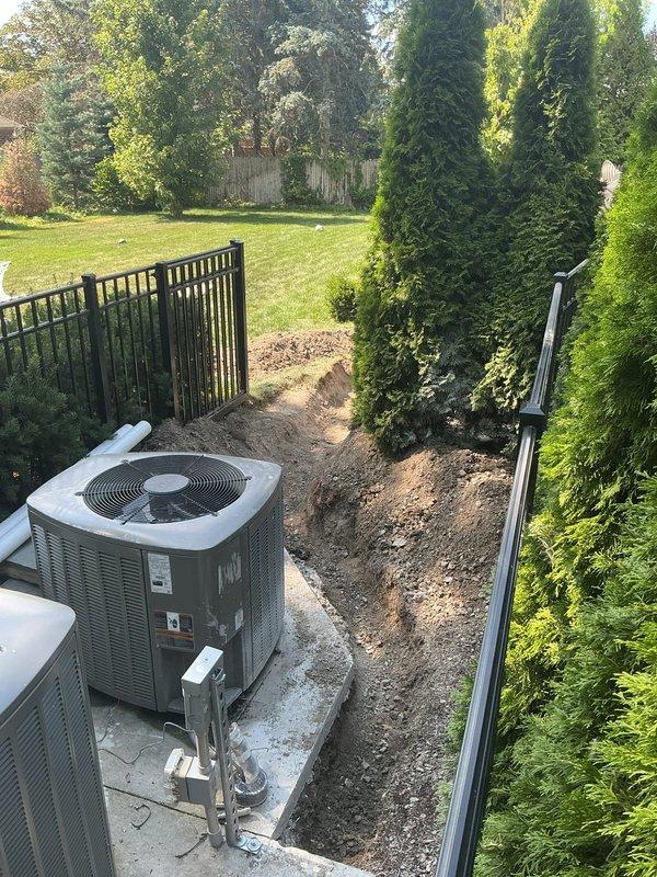 Excavated trench through residential yard following Miss Dig ticket #206072403970 clearance to install underground plumbing line. Trenched from exterior HVAC pad area through landscaped section and across lawn, installed white PVC piping system at proper grade and depth per code requirements, with soil berms maintained along trench edges for backfill.