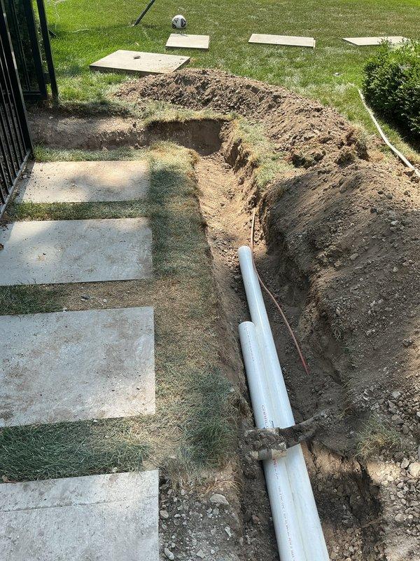 Excavated trench through residential yard following Miss Dig ticket #206072403970 clearance to install underground plumbing line. Trenched from exterior HVAC pad area through landscaped section and across lawn, installed white PVC piping system at proper grade and depth per code requirements, with soil berms maintained along trench edges for backfill.