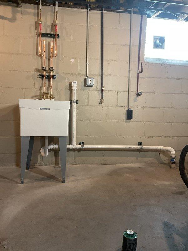 Completed installation of basement plumbing infrastructure including copper supply manifold system with isolation valves, white PVC drainage piping with proper mounting brackets, and utility sink with metal stand. All piping systems were properly secured to concrete block walls, tested for leaks, and verified for proper operation alongside existing HVAC equipment in the mechanical room.