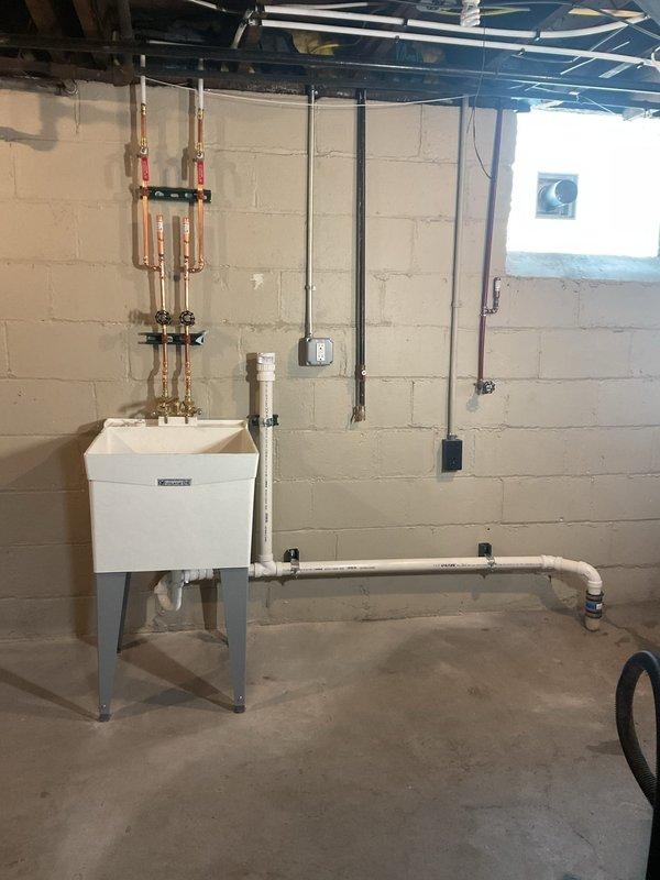Completed installation of basement plumbing infrastructure including copper supply manifold system with isolation valves, white PVC drainage piping with proper mounting brackets, and utility sink with metal stand. All piping systems were properly secured to concrete block walls, tested for leaks, and verified for proper operation alongside existing HVAC equipment in the mechanical room.