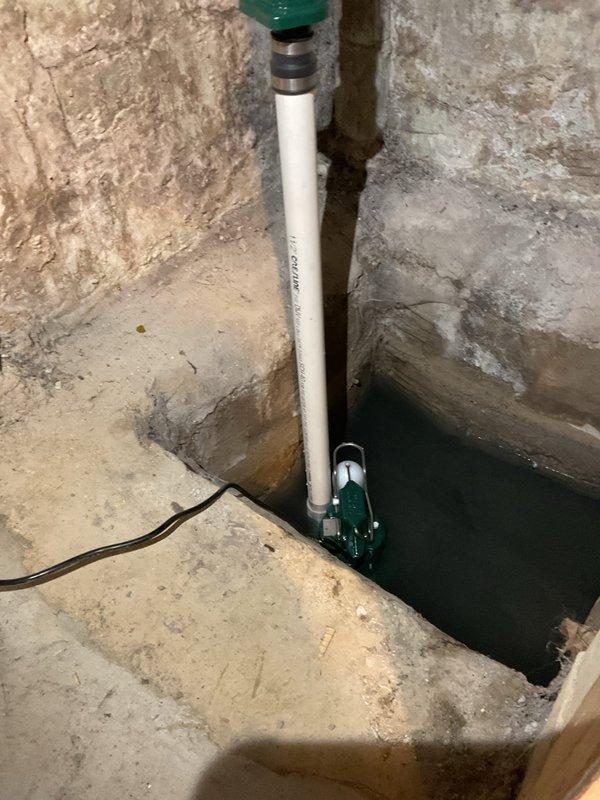 Completed installation of residential sump pump system in basement corner, including green submersible pump unit positioned in sump basin with vertical white PVC discharge piping and green check valve assembly. Routed electrical supply cord across concrete floor and configured discharge line with multiple elbow fittings to direct water flow away from foundation area.