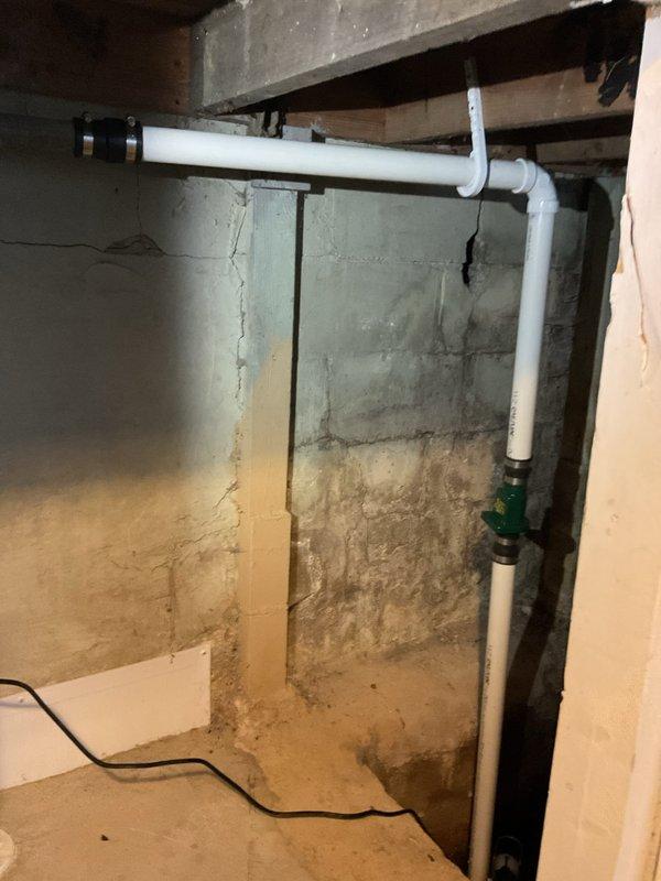 Completed installation of residential sump pump system in basement corner, including green submersible pump unit positioned in sump basin with vertical white PVC discharge piping and green check valve assembly. Routed electrical supply cord across concrete floor and configured discharge line with multiple elbow fittings to direct water flow away from foundation area.