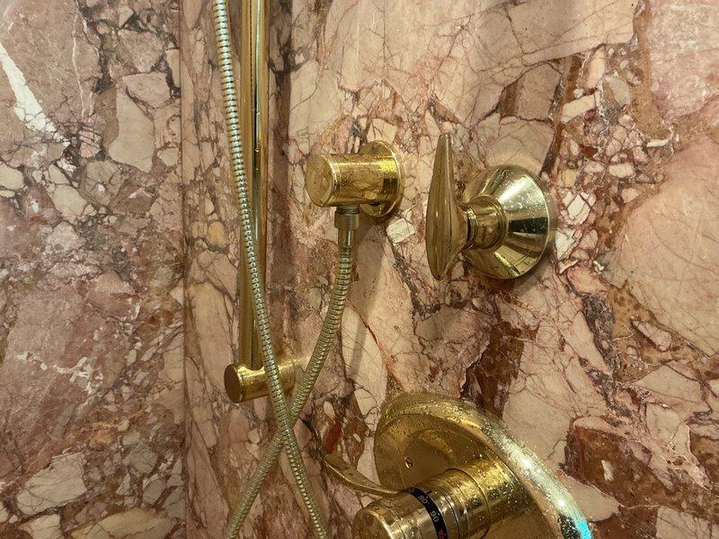 Conducted on-site evaluation of shower fixture assembly mounted on marble wall surface. Assessed brass-finished handheld showerhead system with flexible metal hose, tub spout, and control valve assembly showing visible mineral deposits and water spotting, indicating potential maintenance needs. Documented existing fixture configuration and condition through photographic record for repair estimate preparation.