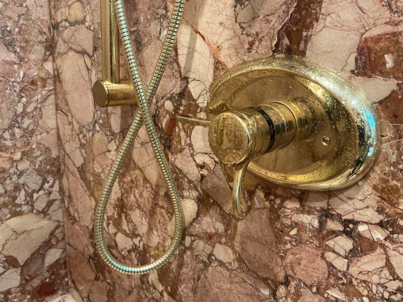 Conducted on-site evaluation of shower fixture assembly mounted on marble wall surface. Assessed brass-finished handheld showerhead system with flexible metal hose, tub spout, and control valve assembly showing visible mineral deposits and water spotting, indicating potential maintenance needs. Documented existing fixture configuration and condition through photographic record for repair estimate preparation.
