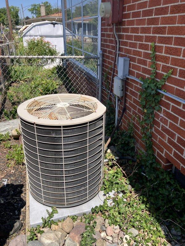 Conducted on-site evaluation of a 20-year-old residential air conditioning system that was blowing air but not cooling. Inspected outdoor condenser unit positioned on concrete pad against brick exterior and assessed attic-mounted air handler equipment. Documented existing system configuration and provided replacement estimate for complete HVAC system due to age and non-cooling condition.