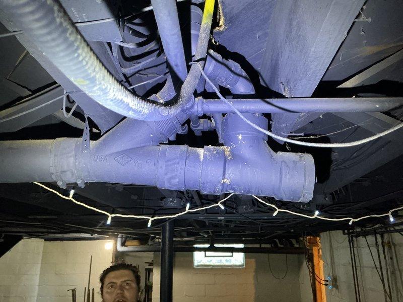 Responded to service call for malfunctioning toilet and drainage issue with waste backing up in pipes. Upon arrival, contacted customer regarding water main break outside property before proceeding with interior plumbing assessment. Investigated toilet malfunction and identified back pitch problem causing waste to accumulate in drainage line. Inspected underfloor plumbing assembly including PVC pipe connections, T-junction fittings, and drainage system configuration in basement utility area. Examined purple PVC piping network with multiple elbows, couplings, and flexible corrugated hoses to diagnose drainage flow issues. Addressed improper pipe slope preventing adequate waste evacuation through the system. Documented existing plumbing configuration and connection points for reference. Resolved toilet operational issues and corrected drainage concerns to restore proper waste flow.