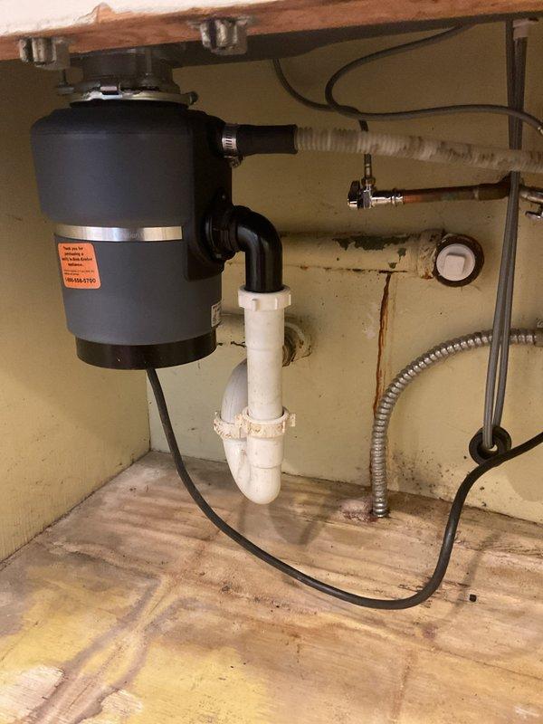 Completed installation of new kitchen sink faucet and garbage disposal unit. Replaced existing fixtures with modern single-handle faucet in brushed nickel finish, mounted on deck plate over stainless steel double-bowl sink configuration. Installed black garbage disposal unit with proper mounting hardware and electrical connection. Connected white PVC drainage system with P-trap configuration to wall drain, ensuring proper slope and secure fittings. Tested all connections for leaks, verified disposal operation, and confirmed water flow through faucet and drainage system functioning properly.