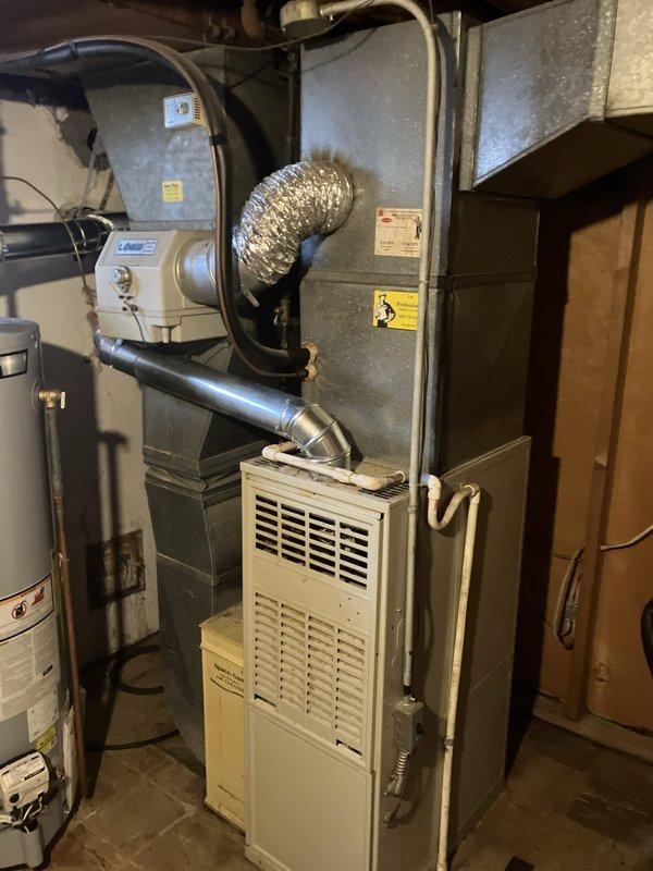 Conducted comprehensive assessment of existing HVAC system for medium-sized residential property requiring full system replacement estimate. Evaluated indoor furnace unit with humidifier and air handler located in basement utility room, along with outdoor cylindrical air conditioning unit positioned against exterior brick wall. Documented current equipment configuration including natural gas furnace, ductwork connections, and aging outdoor condenser showing visible weathering and discoloration. Provided complete replacement estimate for heating and cooling system serving approximately 1500-3000 square feet of conditioned space.