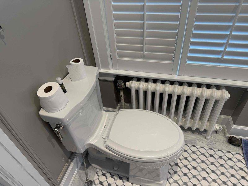 Responded to service call for upstairs bathroom radiator reinstallation assessment. Upon arrival, conducted thorough evaluation of existing radiator system connected to 5-year-old boiler located in basement. Inspected white cast iron multi-column radiator unit and associated plumbing connections, including supply and return piping. Documented current conditions with measurements and photographs showing plumbing fixture assembly, PVC connections, and valve configurations beneath fixture on tiled floor. Assessed radiator mounting requirements, connection points, and compatibility with existing heating system to determine scope of reinstallation work needed.