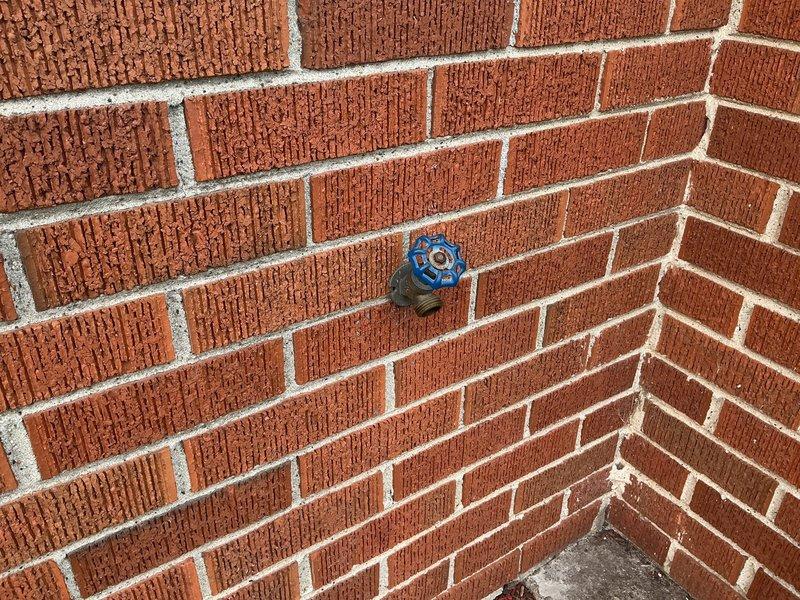 Replaced two exterior hose bibs as specified in the estimate. The first spigot was installed on an exterior brick wall with white PVC stub-out and dark valve handle, while the second featured a brass or copper pipe assembly with blue four-spoke wheel valve handle at an interior brick corner. Both installations were completed with proper mounting and secure connections to existing water supply lines.