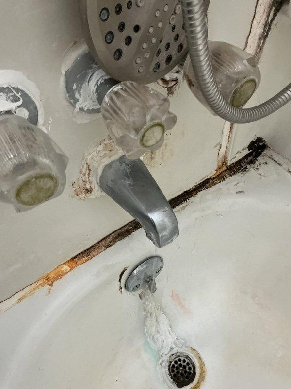 # Bathtub Hot Water Valve Leak Diagnosis and Shower Head Replacement Quote

Responded to service call for leaking hot water valve on bathtub fixture. Performed comprehensive diagnostic inspection of bathroom plumbing system. Identified aging Price Pfister valve assembly with deteriorated internal components causing active leak at hot water control. Inspected surrounding plumbing infrastructure beneath fixture, noting corrosion and water staining on pipe connections and cabinet surfaces consistent with prolonged moisture exposure.

Examined shower and tub area, documenting condition of existing fixtures including chrome showerhead with flexible metal hose attachment, clear plastic valve handles showing age-related discoloration, and tub spout. Noted significant mineral deposits, lime scale buildup on shower hose, and extensive rust staining on tub surface, wall tiles, grout lines, and drain assembly indicating long-term water quality issues and fixture deterioration.

Assessed shower he