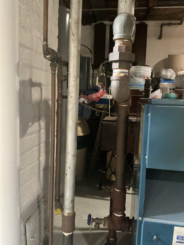 Responded to service call for steam boiler with active leak in return line. Upon inspection, found 25+ year-old steam boiler system in basement with significant corrosion on return piping and fittings. Identified leak source at corroded elbow joint on return line, with visible water staining on concrete floor beneath affected area. Documented extensive oxidation throughout piping system, particularly at connection points and uninsulated sections. Advised homeowner of system condition, age-related deterioration, and recommended replacement of failed return line section and evaluation of overall boiler system integrity due to advanced age and widespread corrosion throughout distribution piping.