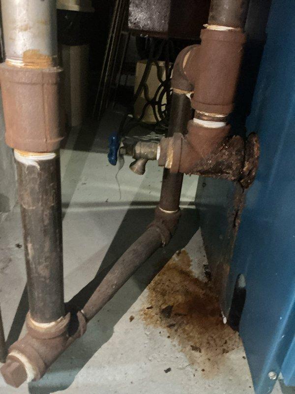 Responded to service call for steam boiler with active leak in return line. Upon inspection, found 25+ year-old steam boiler system in basement with significant corrosion on return piping and fittings. Identified leak source at corroded elbow joint on return line, with visible water staining on concrete floor beneath affected area. Documented extensive oxidation throughout piping system, particularly at connection points and uninsulated sections. Advised homeowner of system condition, age-related deterioration, and recommended replacement of failed return line section and evaluation of overall boiler system integrity due to advanced age and widespread corrosion throughout distribution piping.