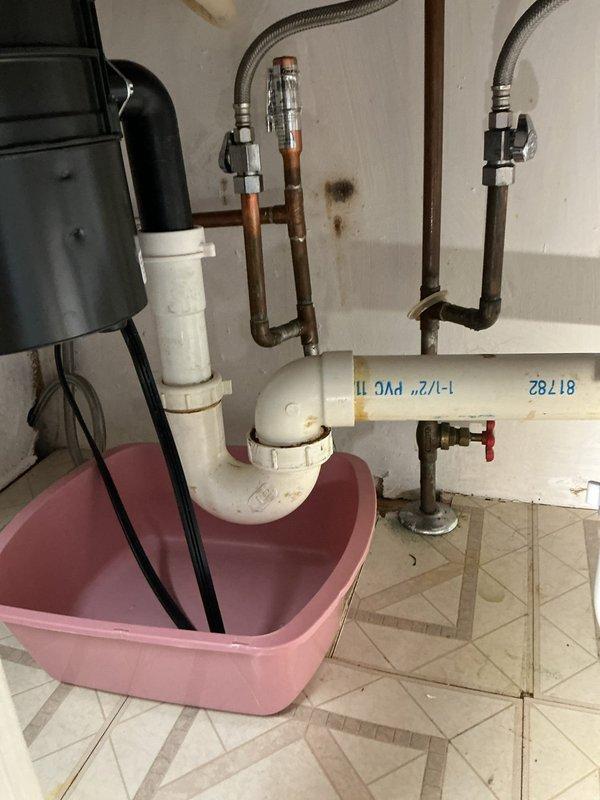 Responded to service call regarding inadequate piping length connecting faucet and garbage disposal, along with cross-contamination issue causing water to run from faucet during shower operation. Upon inspection, discovered improper drain configuration with white PVC piping showing significant internal debris accumulation and blockage. The existing trap assembly was positioned incorrectly with insufficient pipe extensions to reach proper connection points at both the faucet drain and garbage disposal unit.

Documented existing conditions showing copper supply lines with oxidation, flexible braided supply hoses, and compromised PVC drain pipe marked as old installation. The cut section of drain pipe revealed heavy buildup restricting proper water flow, which contributed to the cross-flow problem between fixtures. Verified water heater connections and shut-off valve functionality during assessment. Pink collection pan was positioned beneath trap assembly, indicating previous leak concern