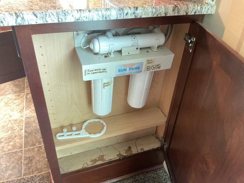 Conducted on-site evaluation of existing SUN PURE under-sink water filtration system for replacement estimate. Assessed current dual-stage filter configuration with vertical cylindrical housings and horizontal mounting assembly, documented plumbing connections and cabinet space constraints. Photographed installation area including P-trap configuration, electrical connections, and cabinet condition to provide accurate replacement system specifications and labor requirements.