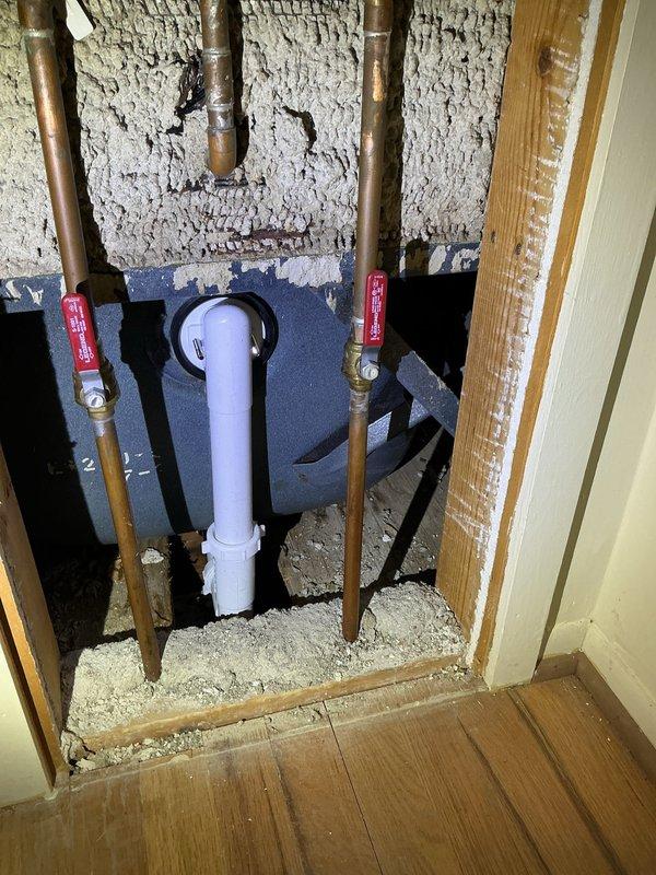 Completed plumbing repair involving replacement of corroded metal drain piping with new PVC drain assembly beneath floor structure. Installed white PVC P-trap configuration with proper joints and connections to replace deteriorated rusted metal pipe that showed significant corrosion. Work included accessing plumbing through exposed floor cavity, connecting new drainage system to existing copper supply lines equipped with red-handled shut-off valves, and ensuring proper drainage flow through the updated PVC piping system.