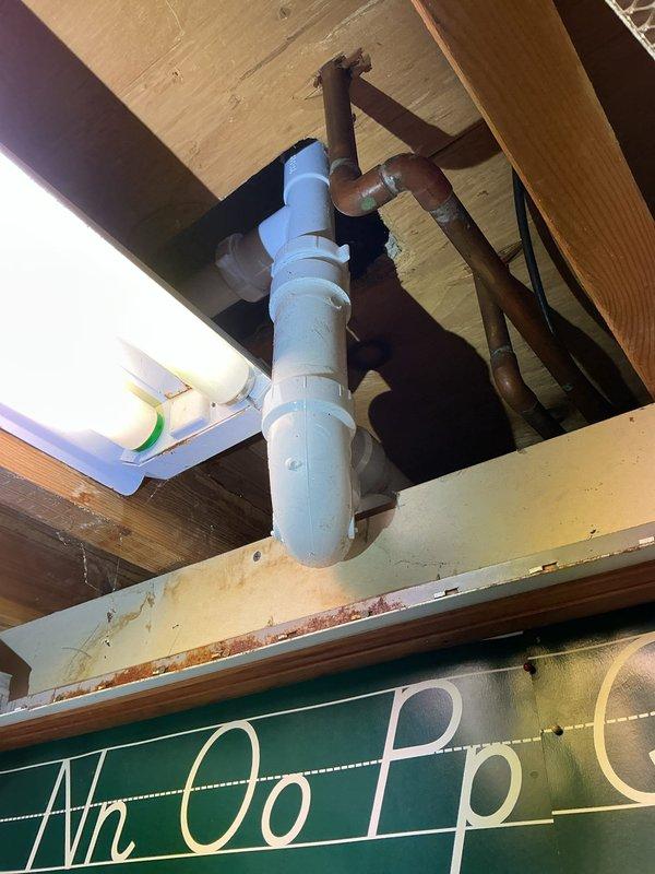 Completed plumbing repair involving replacement of corroded metal drain piping with new PVC drain assembly beneath floor structure. Installed white PVC P-trap configuration with proper joints and connections to replace deteriorated rusted metal pipe that showed significant corrosion. Work included accessing plumbing through exposed floor cavity, connecting new drainage system to existing copper supply lines equipped with red-handled shut-off valves, and ensuring proper drainage flow through the updated PVC piping system.