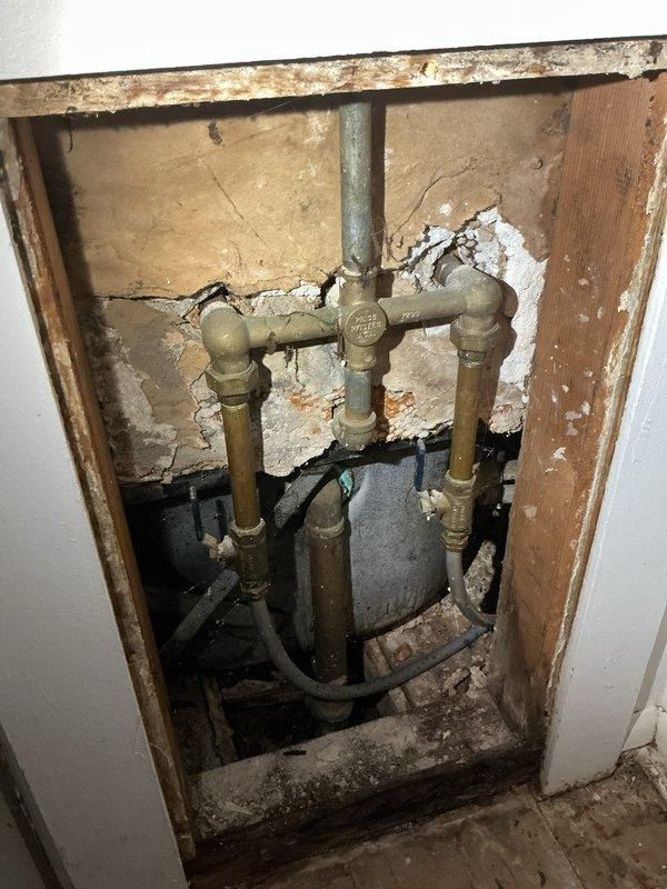 Responded to emergency call for leaking pipe in bathroom of apartment unit. Upon arrival, provided courtesy call 30 minutes prior as requested and accessed the affected area. Inspection revealed aged brass plumbing manifold assembly with multiple connection points showing significant deterioration and active leak. Opened wall cavity to expose full extent of plumbing infrastructure, discovering tarnished brass/bronze pipes with compromised joints and fittings. Wall backing exhibited water staining and extensive moisture damage, indicating prolonged leak condition. 

Assessed lower plumbing connections including black flexible tubing and dark-colored components at base of assembly. Further investigation revealed additional aged piping in crawl space/basement area with visible corrosion and patina on brass-toned pipe. Wooden framing and structural elements showed water damage and deterioration from moisture exposure. Documented existing conditions through photographs showing compromised d