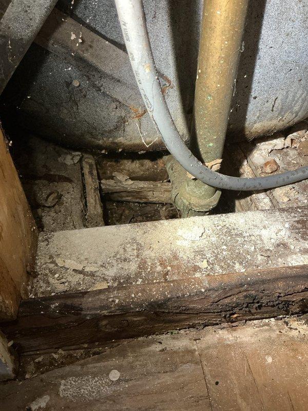 Responded to emergency call for leaking pipe in bathroom of apartment unit. Upon arrival, provided courtesy call 30 minutes prior as requested and accessed the affected area. Inspection revealed aged brass plumbing manifold assembly with multiple connection points showing significant deterioration and active leak. Opened wall cavity to expose full extent of plumbing infrastructure, discovering tarnished brass/bronze pipes with compromised joints and fittings. Wall backing exhibited water staining and extensive moisture damage, indicating prolonged leak condition. 

Assessed lower plumbing connections including black flexible tubing and dark-colored components at base of assembly. Further investigation revealed additional aged piping in crawl space/basement area with visible corrosion and patina on brass-toned pipe. Wooden framing and structural elements showed water damage and deterioration from moisture exposure. Documented existing conditions through photographs showing compromised d