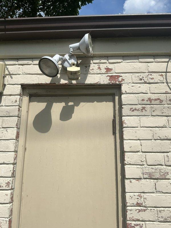 Installed dual outdoor motion-activated security floodlight fixture on exterior white brick wall above building entrance. Mounted white adjustable twin-head fixture to wall, completed electrical connections, and tested motion sensor functionality. Verified proper operation of both floodlight heads and confirmed motion detection coverage for yard area as requested.