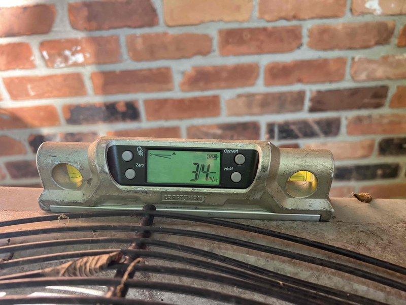 Responded to service call for outdoor condensing unit evaluation. Upon arrival, confirmed unit bears flame stickers indicating previous installation, though installer verification was requested. Conducted level assessment of approximately 2007-vintage air conditioning condenser positioned on concrete pad in basement area. Using precision digital level instrument, measured unit alignment and documented 74°F ambient temperature reading during inspection. Observed condensing unit sitting on concrete base with surrounding ground cover including rocks, soil, and vegetation encroachment. Photographically documented current installation conditions, including unit positioning, concrete pad status, and level measurements. Confirmed condenser is not properly leveled on existing pad. Provided assessment findings and discussed restoration options to properly level equipment for optimal performance and longevity.