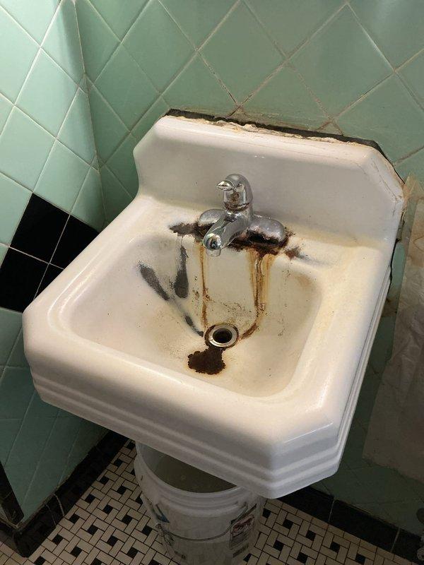 Removed and replaced deteriorated pedestal sink and faucet assembly per customer estimate. The existing wall-mounted sink exhibited extensive rust staining, corrosion around fixtures, and damaged porcelain along the mounting edge. Disconnected water supply lines and removed old chrome-plated faucet with separate hot and cold handles. Detached compromised sink from wall mounting, ensuring proper care around vintage mint green diagonal tile work and black tile wainscoting. Installed new pedestal sink unit with updated chrome faucet assembly. Connected hot and cold water supply lines, tested for leaks, and verified proper drainage. All fixtures secured and operational upon completion.