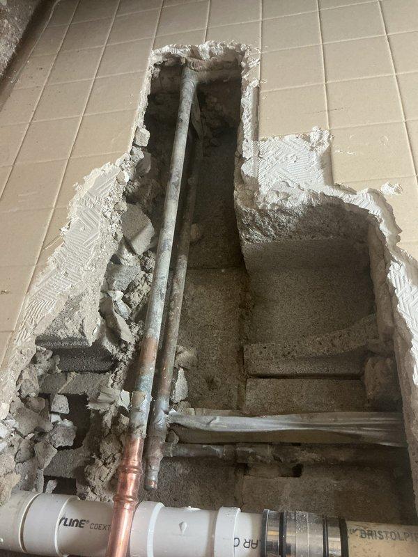 Performed exploratory wall and floor openings to access and assess concealed plumbing system in commercial facility. Cut rectangular access points through beige tile floor and wall surfaces, exposing underlying pipe infrastructure including white PVC drainage lines, copper supply piping, and various fittings and connections. Identified and evaluated condition of existing plumbing components within wall cavity and floor substrate for diagnostic purposes related to sold repair work.