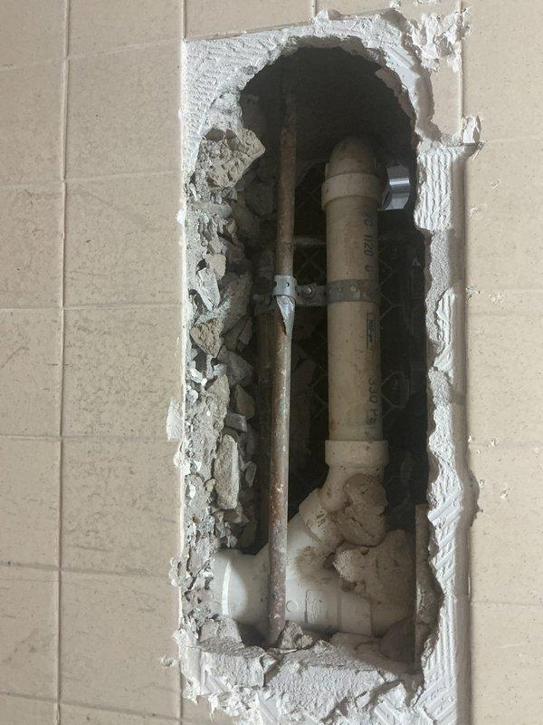 Performed exploratory wall and floor openings to access and assess concealed plumbing system in commercial facility. Cut rectangular access points through beige tile floor and wall surfaces, exposing underlying pipe infrastructure including white PVC drainage lines, copper supply piping, and various fittings and connections. Identified and evaluated condition of existing plumbing components within wall cavity and floor substrate for diagnostic purposes related to sold repair work.