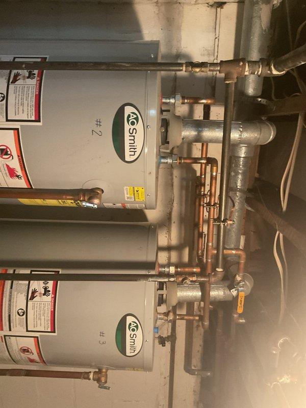 Responded to service call for insufficient hot water production from tanked water heater system. Accessed basement utility room through tenant units to inspect dual A.O. Smith water heater installation consisting of units #2 and #3. Performed diagnostic evaluation of both tanked water heaters, examining gas control valves, copper supply and distribution piping, shut-off valves, and overall system configuration. Inspected manufacturer labels, safety warnings, and operational specifications on both units. Assessed piping connections, joints, elbows, and valve assemblies for proper function. Evaluated gas supply system and burner operation to determine cause of inadequate heating capacity. Documented existing installation condition including copper piping patina, insulated pipe runs, and dual-unit configuration designed for high-capacity residential hot water demand.