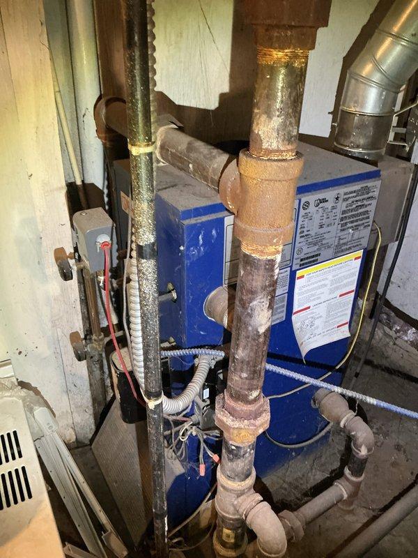 Responded to service call for malfunctioning radiator steam control valve on 6-year-old steam boiler system located in basement. Upon arrival, wore protective shoe covers per customer request due to new carpeting. Inspected blue boiler unit and associated piping system, noting considerable corrosion and oxidation on copper piping and brass fittings throughout the installation. Examined steam distribution system including multiple vertical risers, control valves, and connections. Evaluated the problematic radiator valve controlling steam flow and assessed overall system condition. Documented equipment showing signs of age-related deterioration including corroded piping, worn insulation, and oxidized fittings. Photographed boiler unit with specification labels, sight glasses, control components, and surrounding mechanical connections for service records.