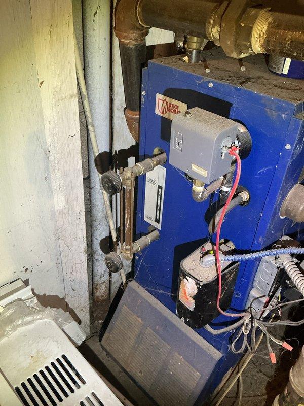 Responded to service call for malfunctioning radiator steam control valve on 6-year-old steam boiler system located in basement. Upon arrival, wore protective shoe covers per customer request due to new carpeting. Inspected blue boiler unit and associated piping system, noting considerable corrosion and oxidation on copper piping and brass fittings throughout the installation. Examined steam distribution system including multiple vertical risers, control valves, and connections. Evaluated the problematic radiator valve controlling steam flow and assessed overall system condition. Documented equipment showing signs of age-related deterioration including corroded piping, worn insulation, and oxidized fittings. Photographed boiler unit with specification labels, sight glasses, control components, and surrounding mechanical connections for service records.