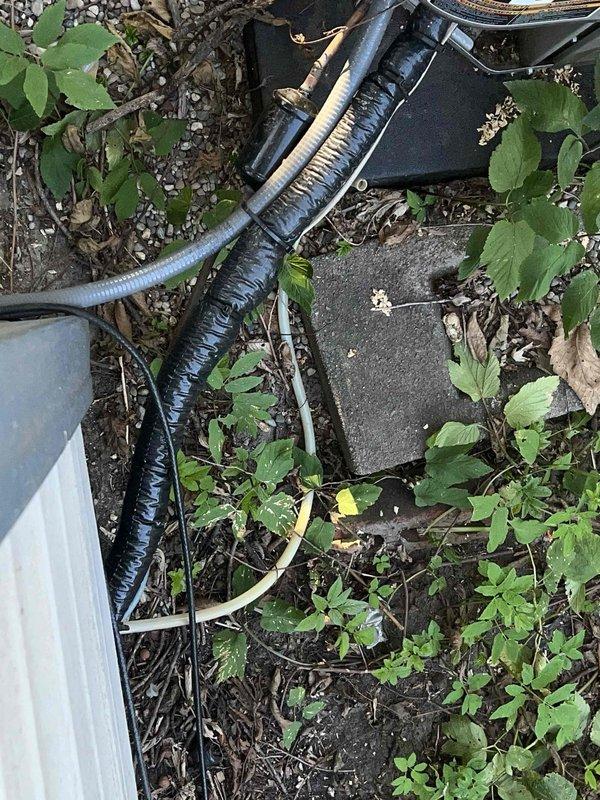 Responded to service call for water leaking from bottom of furnace causing wet floors in encapsulated crawl space. Upon inspection, found recently installed furnace and AC system with unit not shutting off properly, though system was still providing cooling. Identified water intrusion originating from underneath the unit area. Inspected exterior condensate drainage system and flexible discharge line routing. Photographed conditions including crawl space access point and exterior mechanical components with drainage piping. Documented weathered deck structure near unit location and condensate line pathway through vegetation and ground-level area. System evaluated for proper drainage function and potential sources of water accumulation beneath furnace installation.