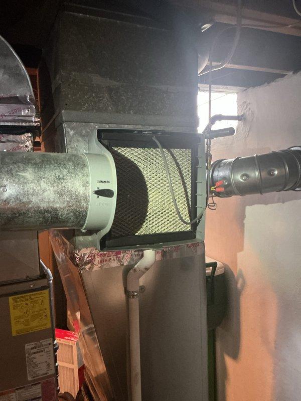 Completed comprehensive Gold AC inspection on 9-year-old air conditioning system and 3-year-old furnace located in basement. Performed full system evaluation including thermostat functionality check, verified White Rodgers digital thermostat displaying proper temperature readings with system operating normally. Inspected furnace unit, blower assembly, and air filtration system; noted heavy dust accumulation on cylindrical blower housing requiring attention during future maintenance. Verified air filter condition in black frame housing, checked flexible ductwork connections, and assessed overall system performance with all equipment functioning as designed at time of inspection.