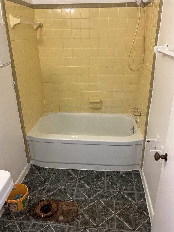 Completed bathtub replacement in commercial bathroom per approved estimate. Removed existing deteriorated bathtub and corroded plumbing fixtures, including heavily rusted flange components. Installed new white bathtub with integrated front panel against cream-tiled walls. Mounted chrome shower fixture and handheld showerhead assembly with hose. Connected brass-toned faucet fixtures on tub deck. Configured under-tub plumbing access panel with hot and cold water supply lines (marked with red and blue caps), copper piping, and white PVC P-trap drain assembly with proper fittings. Secured access panel with wooden trim and mounting hardware. Verified all connections for proper operation and leak-free performance. New bathtub picked up from Advanced Plumbing supply house per purchase order #1366314.